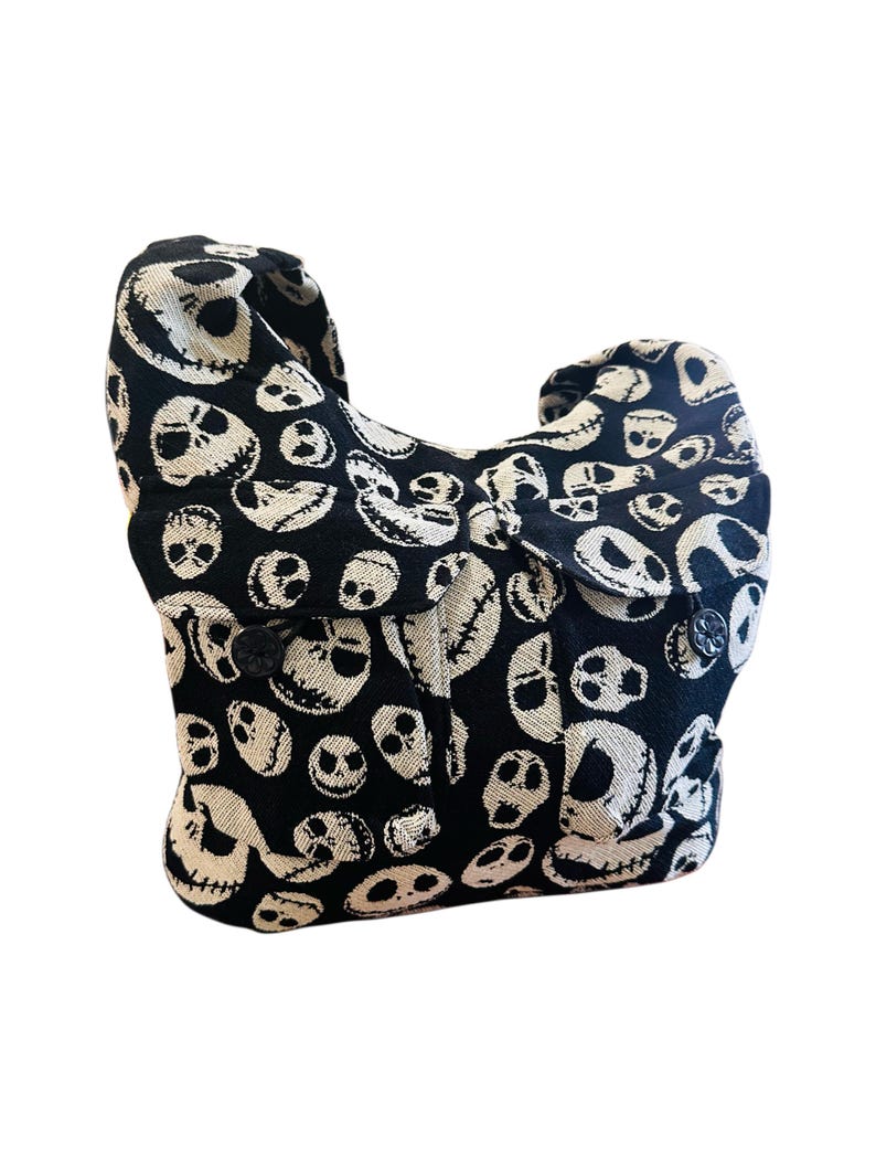 May include: A black and off-white hobo bag featuring an all-over woven pattern of various skull faces with stitched-style mouths. The bag has a single shoulder strap, a slouchy silhouette, and two front pockets secured with black flower-shaped buttons.