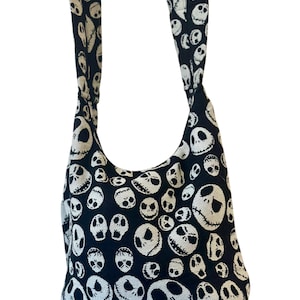 May include: A black hobo-style shoulder bag featuring an all-over woven pattern of white skull faces with stitched-style mouths and hollow eyes. The bag includes a wide, integrated shoulder strap made from the same fabric as the main body.