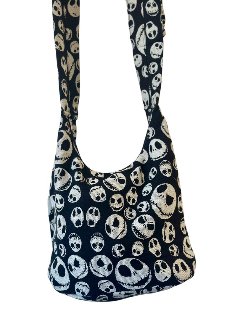 May include: A black hobo-style shoulder bag featuring an all-over woven pattern of white skull faces with stitched-style mouths and hollow eyes. The bag includes a wide, integrated shoulder strap made from the same fabric as the main body.