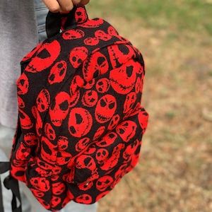 May include: Black and red backpack featuring a repeating pattern of stylized skull faces. The backpack has a top handle and adjustable shoulder straps. The design is a bold contrast of black and red, creating a striking visual effect.