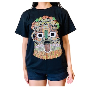 May include: Black t-shirt featuring a colorful, geometric mask design. The mask has a detailed pattern with orange, green, yellow, and red accents. The design includes eye shapes and a fringed bottom. The shirt is worn by a person.