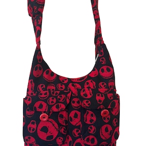 May include: A black crossbody bag featuring a repeating red skull pattern. The bag includes a long shoulder strap with a red button detail, two front pockets with flap closures, and two prominent red buttons securing the front pocket flaps.