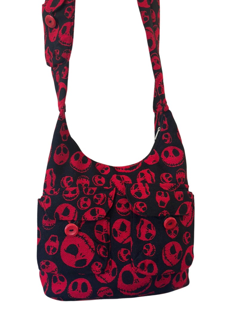 May include: A black crossbody bag featuring a repeating red skull pattern. The bag includes a long shoulder strap with a red button detail, two front pockets with flap closures, and two prominent red buttons securing the front pocket flaps.