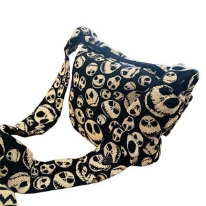 May include: A black and cream-coloured crossbody bag featuring an all-over woven pattern of various skull faces with stitched-style mouths and hollow eyes. The bag includes a wide, matching fabric shoulder strap and a zippered top closure.