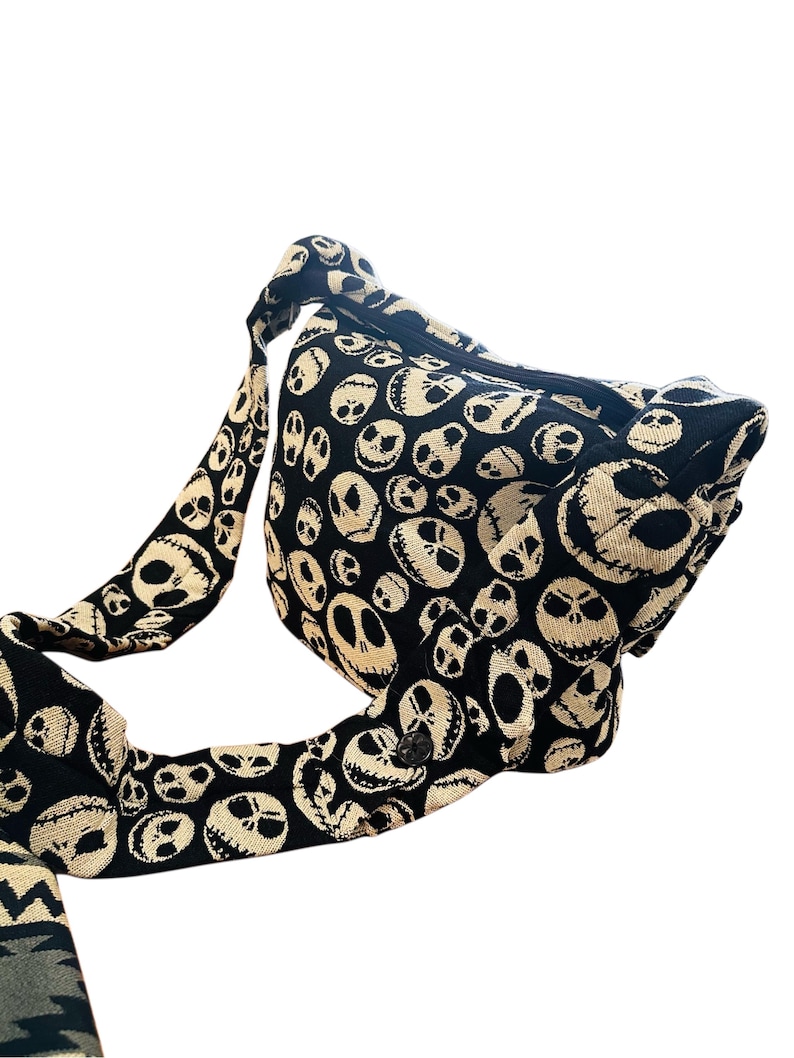 May include: A black and cream-coloured crossbody bag featuring an all-over woven pattern of various skull faces with stitched-style mouths and hollow eyes. The bag includes a wide, matching fabric shoulder strap and a zippered top closure.