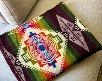 May include: A folded blanket with a geometric design in shades of red, orange, green, and purple. The central pattern is a circular design with a blue center, surrounded by a white background and framed by a diamond shape.