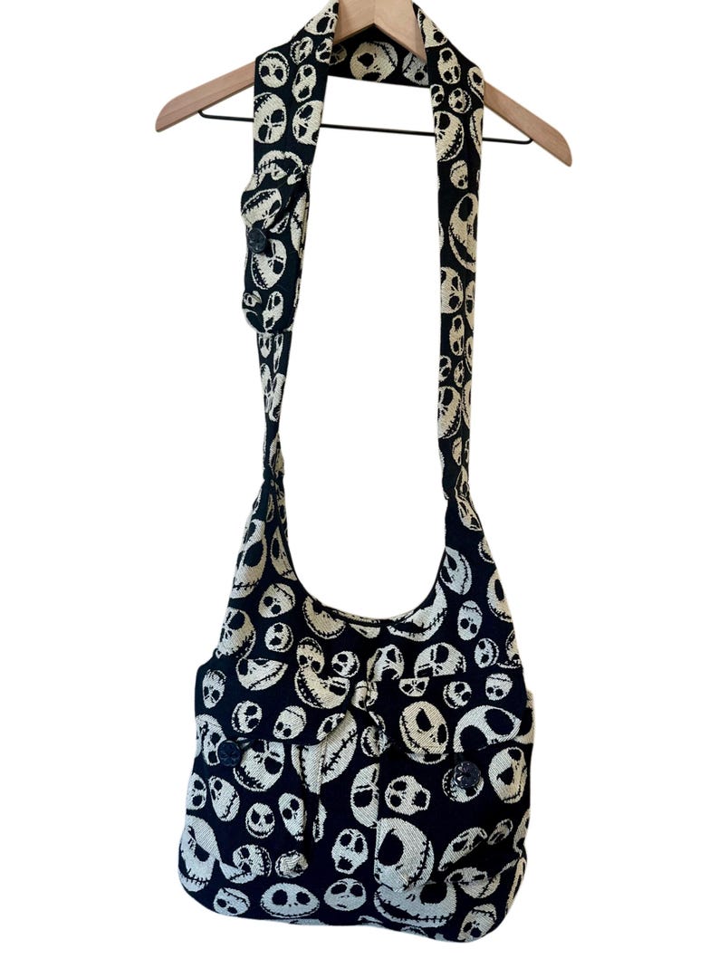 May include: A black and off-white hobo-style shoulder bag featuring an all-over pattern of stylised skull faces. The bag includes a long, wide shoulder strap and two front pockets with button closures, all crafted from a woven fabric material.