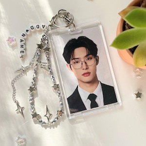 May include: A clear plastic ID holder with a photo of a person wearing glasses and a black suit. Attached is a silver beaded chain with star charms and the word "NAME" spelled out in white letter beads. Decorative star-shaped accents are scattered around.
