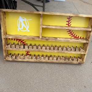 Könnte beinhalten: Baseball-Display-Holzkiste mit gelbem Hintergrund und Baseball-Naht-Design. Die Kiste hat mehrere Regale und Stifte zum Halten von Basebällen. Eine weiße Baseballspieler-Silhouette befindet sich oben links.