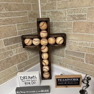 Puede incluir: Una cruz de madera marrón oscuro llena de pelotas de béisbol, colocada contra una pared de ladrillos. La imagen también muestra el texto "DREAMS don't work unless YOU DO", "TEAMWORK makes the dream work", y "ALL things are possible if you BELIEVE".