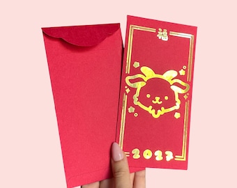 May include: Two rectangular red paper envelopes. One envelope is plain, while the other features a gold foil design of a goat head, the Chinese character for fortune, and the year 2023, all enclosed within a thin gold rectangular border.