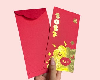 May include: Two red paper envelopes. One envelope is plain with a scalloped flap. The other features gold foil text reading 2024, a gold Chinese character, and a gold foil illustration of a stylised sheep with a smiling face.