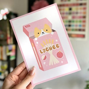 May include: A pink and white art print featuring a cartoon illustration of a box of lychee-flavored biscuit sticks. The box has a corgi peeking out. The word "Lychee" is written in brown letters. The print is held by a hand.