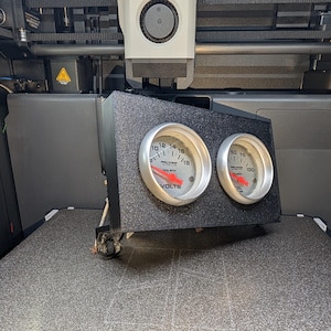 May include: A black textured panel with two circular gauges. The left gauge reads "VOLTS" and the right gauge reads "PSI". Both gauges have silver rims, white faces, and red needles. The panel is mounted in a vehicle interior.