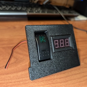 Puede incluir: Panel electrónico negro con un interruptor basculante y una pantalla digital que muestra "8.8.8". El interruptor tiene un indicador luminoso verde. Cables rojos y negros se extienden desde el panel.