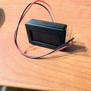 May include: A black electronic component with a rectangular display and red and black wires extending from it. The device is on a wooden surface, with the wires curving away from the component.