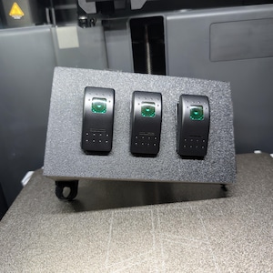 Puede incluir: Un panel gris con tres interruptores basculantes negros, cada uno con un indicador luminoso verde. El panel es probablemente para uso automotriz o industrial. Los interruptores están espaciados uniformemente.