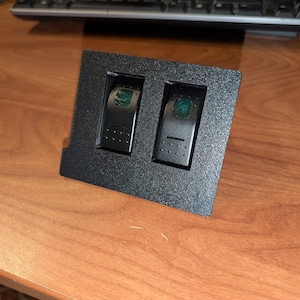 May include: A black, rectangular panel with two rocker switches. Each switch has a black surface with a green light. The panel is set on a wooden surface, with a keyboard and screwdriver visible in the background.