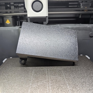Könnte beinhalten: Nahaufnahme eines 3D-Druckers in Betrieb, mit einem teilweise gedruckten rechteckigen Objekt im Vordergrund. Das Objekt ist dunkelgrau und hat eine strukturierte Oberfläche. Die Komponenten des Druckers sind im Hintergrund sichtbar.