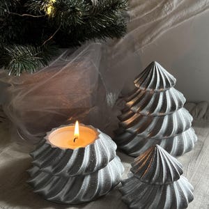 May include: Three silver, glittery, tree-shaped objects with a spiraled, tiered design. One object is a candle holder with a lit, white wax candle inside. The other two objects are solid, decorative tree-shaped figures of varying sizes.