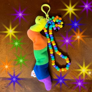 May include: A colorful plush worm keychain with a beaded lanyard. The worm is segmented in rainbow colors, with a yellow head and purple foot. The lanyard features a mix of colorful beads and a yellow clip.