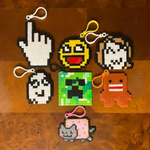 May include: A collection of pixel art keychains featuring various designs. Included are a white cursor, a yellow smiley face, a pixelated character, a white ghost, a green Creeper, an orange Domo, and a Nyan Cat.