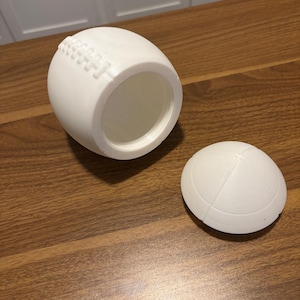 May include: A white, football-shaped container with a removable lid. The container has a textured surface that mimics the stitching of a football. The lid is also football-shaped. The container is sitting on a wooden surface.