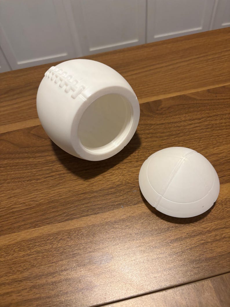 May include: A white, football-shaped container with a removable lid. The container has a textured surface that mimics the stitching of a football. The lid is also football-shaped. The container is sitting on a wooden surface.