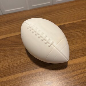 May include: A white football with detailed stitching and a textured surface. The football is oval-shaped and rests on a wooden surface. The design includes the characteristic laces and panels of a standard football.