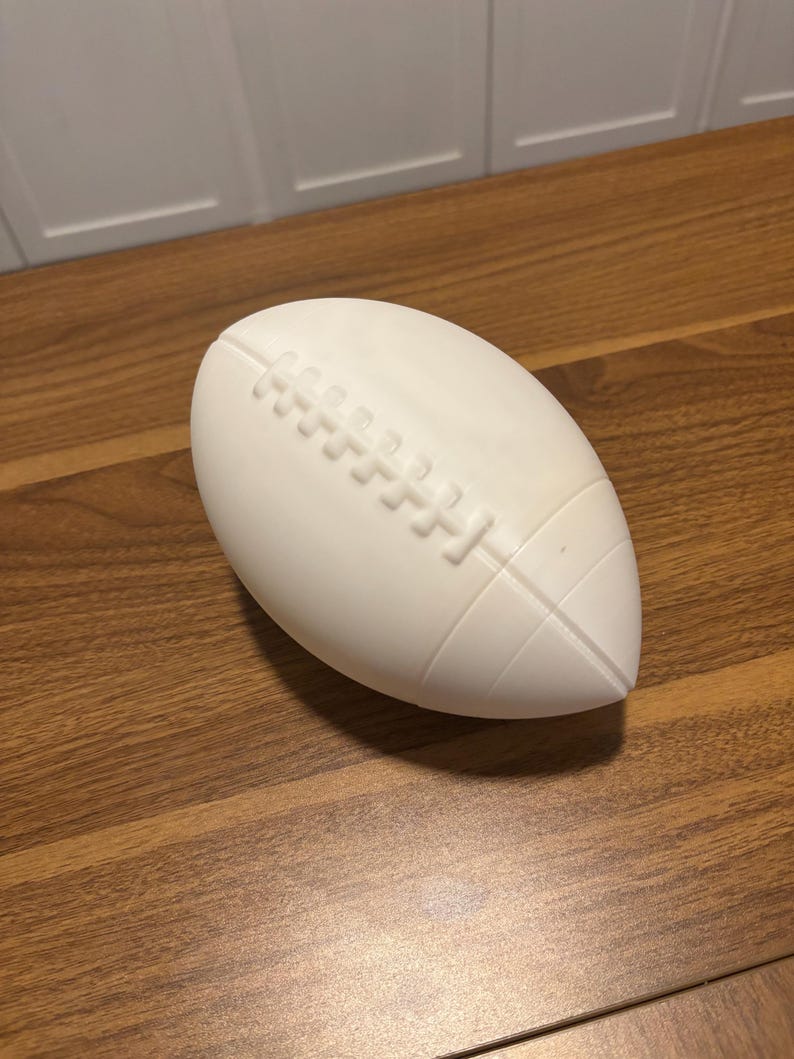 May include: A white football with detailed stitching and a textured surface. The football is oval-shaped and rests on a wooden surface. The design includes the characteristic laces and panels of a standard football.
