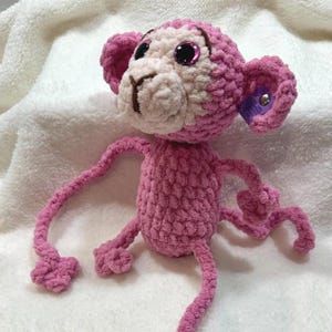 Puede incluir: Una muñeca de mono rosa de ganchillo hecha a mano con brazos y piernas largos. El mono tiene una cara blanca, orejas rosas y ojos negros grandes y brillantes. La muñeca está hecha de hilo suave y texturizado.
