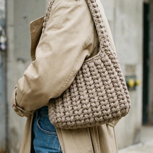 May include: A beige, hand-knitted shoulder bag with a thick, textured stitch pattern. The bag has a rounded shape and a wide strap. The bag is carried over the shoulder of a person wearing a tan trench coat and blue jeans.