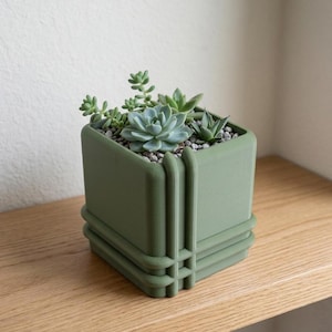 May include: A square, olive green planter filled with various succulents and small gray pebbles. The planter has a geometric design with vertical and horizontal lines. The planter sits on a light brown wooden shelf against a white wall.