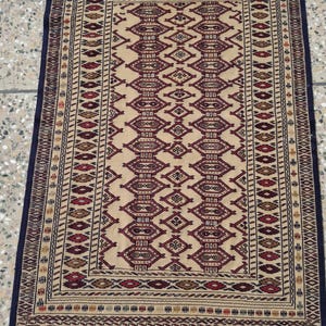 May include: A rectangular woven rug featuring a beige background with a central column of repeating diamond-shaped geometric motifs in deep red, brown, and black. The border displays intricate linear patterns, and the ends have short fringe.