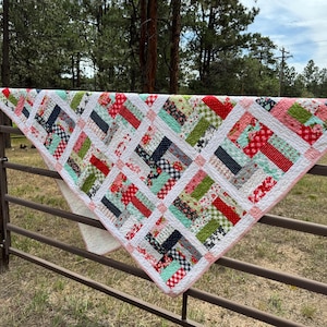 May include: A colorful quilt draped over a metal fence. The quilt features a patchwork design with red, white, green, and navy blue fabrics. The quilt has a white border with a pink trim. The background includes trees and a blue sky.
