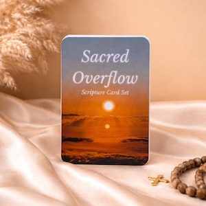 May include: A card set titled "Sacred Overflow Scripture Card Set" with a sunset image. The card is on a satin surface with a wooden bead necklace and a gold cross.