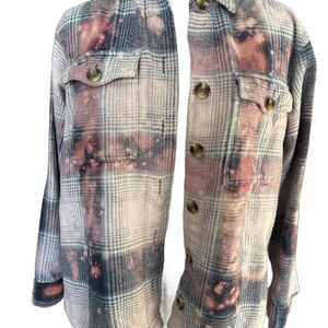 May include: A plaid button-up shirt in shades of pink, gray, and navy blue, with a bleached effect. The shirt has two chest pockets and brown buttons. The long-sleeved shirt is displayed on a mannequin.