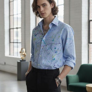 May include: A light blue long-sleeved button-down shirt featuring a multi-coloured paint splatter pattern in shades of purple, teal, and lime green. The shirt has a classic collar, white buttons, and is tucked into a pair of solid black trousers.
