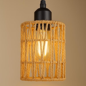 Peut inclure: Un luminaire suspendu avec un abat-jour cylindrique en corde tressée. L'abat-jour entoure une ampoule allumée. Le luminaire a un dessus en métal noir et un seul cordon de suspension. Le luminaire est de couleur chaude et neutre.
