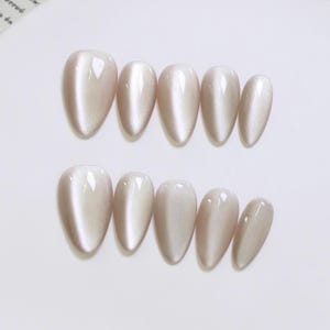 May include: A set of ten almond-shaped, light beige press-on nails. The nails have a glossy finish with a subtle shimmer, arranged in two rows of five on a white surface. The overall effect is elegant and neutral.