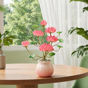 以下が含まれることがあります： 層状の花びらを持つ鮮やかなピンクの蓮の花と、細い質感のある茎についた小さな緑の葉が特徴の装飾用人工植物。植物は、小さな白い小石で満たされた滑らかな淡いピンクのセラミック製の花瓶に植えられています。