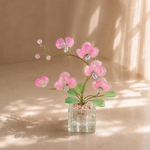 以下が含まれることがあります： 半透明のピンクの花びらと虹色に輝く中心部、緑の葉、小さなピンクの蕾を持つ、ワイヤーで彫刻された装飾用の蘭の植物。この植物は、小さな明るい色の装飾石で満たされた透明な正方形のガラスの花瓶に植えられています。