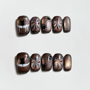 May include: A set of press-on nails with a brown glitter base. Designs include a black cat silhouette, paw prints, and colorful firework-like patterns. The nails are oval-shaped and vary in design.