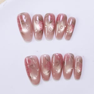 以下が含まれることがあります： 淡いピンク色の人工爪のセット。ピンクと茶色の繊細なバラのデザインで飾られています。各爪は光沢のある仕上がりで、小さな透明な水滴のアクセントがあり、女性的でエレガントな美しさを生み出しています。