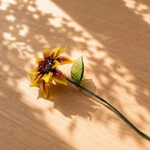 以下が含まれることがあります： 鮮やかな黄色の花びらと根元の濃い赤のアクセント、濃い茶色の中心部、そして細い緑色のガラスの茎に付いた一枚の緑色のガラスの葉が特徴の、手作りのガラス製ヒマワリの彫刻です。精巧な花の造形が施されています。