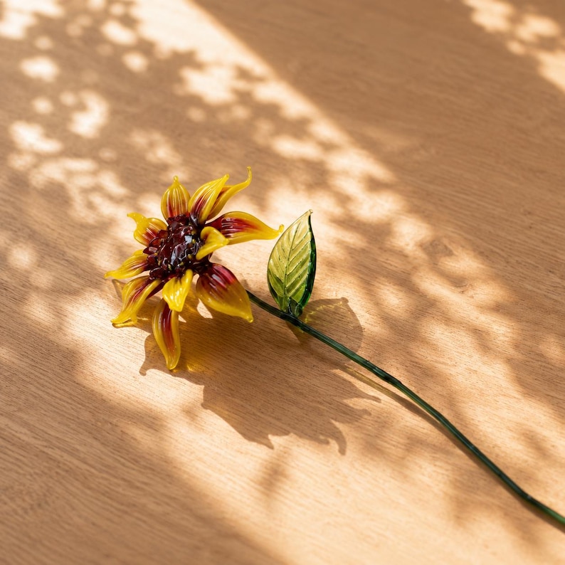 以下が含まれることがあります： 鮮やかな黄色の花びらと根元の濃い赤のアクセント、濃い茶色の中心部、そして細い緑色のガラスの茎に付いた一枚の緑色のガラスの葉が特徴の、手作りのガラス製ヒマワリの彫刻です。精巧な花の造形が施されています。