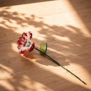 以下が含まれることがあります： 白い花びらの縁が濃い赤色に彩られた、フリル状の花びらを持つガラス製の花の彫刻。花は細い濃い緑色のガラスの茎につながっており、茎の側面には質感のある明るい緑色の葉が1枚付いています。