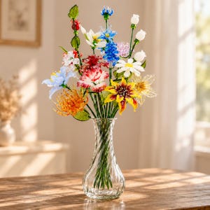 以下が含まれることがあります： 螺旋状の畝模様が施された透明なガラスの花瓶に、色とりどりのガラス製の花束が生けられています。赤、黄、青、白、オレンジの色合いの様々な花が、緑色のガラスの茎と葉と共に配置されています。