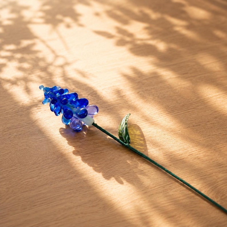 以下が含まれることがあります： 小さな丸い青と紫のガラスビーズの房が先端に付いたムスカリの花のガラス彫刻。細く真っ直ぐな緑色のガラスの茎に、尖った緑色のガラスの葉が1枚横から伸びているデザインで、全体が繊細なガラス細工で構成されています。