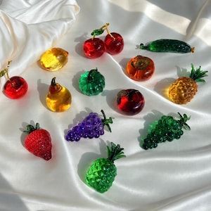 Könnte beinhalten: Eine Sammlung von Miniatur-Glasfiguren in Form von Früchten, darunter rote Kirschen, eine gelbe Zitrone, eine grüne Gurke, eine orangefarbene Kaki, eine gelbe Birne, ein grüner Kürbis, eine Ananas, eine rote Erdbeere sowie violette und grüne Weintrauben.